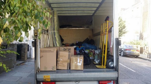 The image shows the open rear of a delivery van parked on a residential street with pavement and parked cars visible on the right side. Inside the van, there is a mix of various waste items and packaging materials, including several cardboard boxes, some flattened and others unopened, with printed labels and logos visible on a few of them. Also present are black plastic bags filled with waste, a yellow hand truck leaning against the side of the van, and a small blue trolley or bin partially visible behind the boxes. The interior of the van has a plain, light-colored lining with a dark ceiling, and some additional miscellaneous items can be seen towards the back. The environment around the van appears to be a quiet suburban area, with a leafy tree on the left and residential buildings partially visible in the background, subtly tying into local waste management services in Honor Oak.