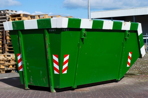 A large green skip bin used for rubbish removal, situated on a paved area possibly near commercial or industrial premises. The bin features a durable metal construction with a smooth surface, painted in bright green with reflective red-and-white chevron stripes on the sides for visibility. It has a wide front opening with a hinged lid that is currently closed, and structural support beams visible along the edges. In the background, there are stacked wooden pallets on the left and a metal-clad building on the right, set against a clear sky. The skip's position and design suggest it is intended for collecting waste materials during commercial waste clearance operations, typical of services provided by Commercial Waste Honor Oak, serving the local community in the Honor Oak area, London postcode. The scene appears to be part of a waste management or rubbish collection site, emphasizing professional rubbish removal in the local area.