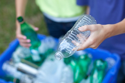 A close-up of a person's hand holding a clear plastic bottle with a slightly ridged surface, empty and without a label, positioned in the foreground. In the background, another individual, partially visible, is standing behind a blue recycling bin filled with various recyclable plastic bottles and containers, some green and some transparent, along with other mixed waste. The setting appears to be outdoors, possibly on a pavement or driveway, with the focus on waste collection and recycling activities associated with rubbish removal services like those provided by Commercial Waste Honor Oak. The scene emphasizes the careful handling of recyclable plastics during rubbish clearance, supporting greener waste management practices in the Honor Oak area.