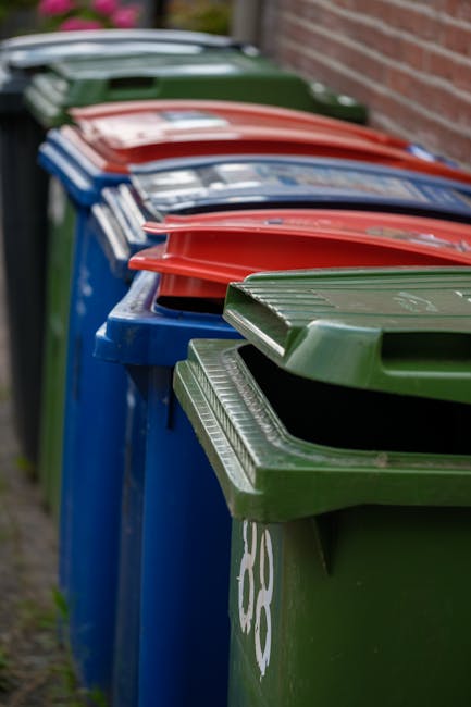 A person's hand is holding a green and blue plastic rubbish bag, grasped at the tied top, with the bag appearing semi-transparent and filled with waste. The bag is positioned centrally in the image ag